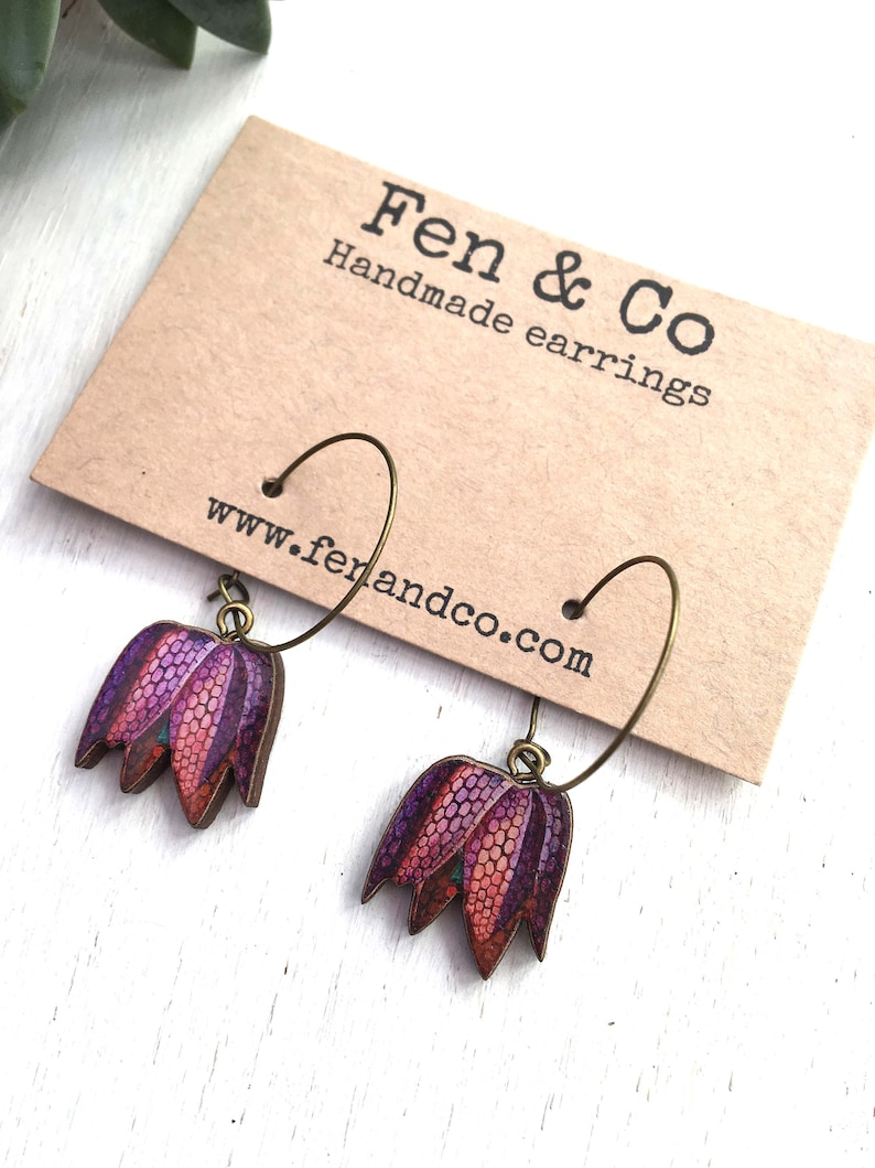 May include: A pair of handmade earrings featuring a purple and orange flower design. The earrings are made of wood and have a bronze hoop.