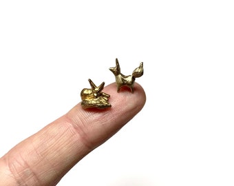 May include: Two small gold fox figurines, one standing and one lying down, are shown on a finger.