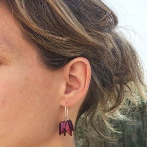 May include: A pair of silver hoop earrings with a pink and purple flower charm. The flower charm has a textured surface and is made of metal.
