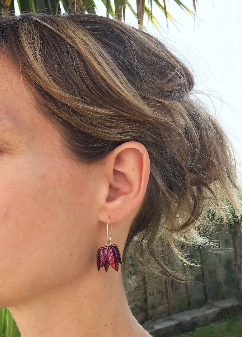 May include: A pair of silver hoop earrings with a pink and purple flower charm. The flower charm has a textured surface and is made of metal.