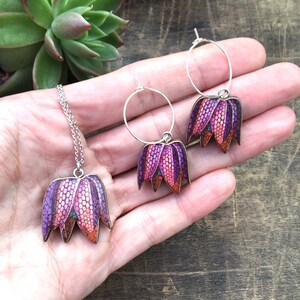 May include: A set of jewelry featuring a necklace and earrings with a floral design. The pendant and earrings are shaped like a flower with purple, pink, and orange petals. The jewelry is made of metal and has a silver finish.