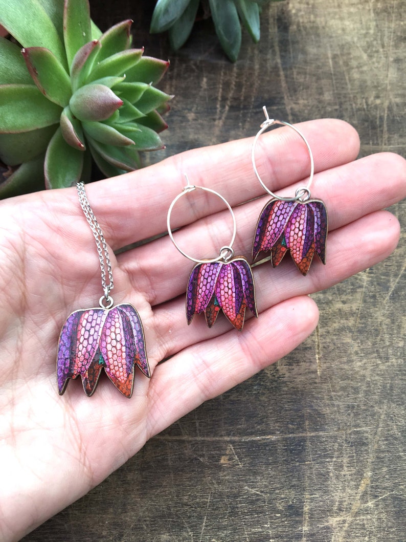 May include: A set of jewelry featuring a necklace and earrings with a floral design. The pendant and earrings are shaped like a flower with purple, pink, and orange petals. The jewelry is made of metal and has a silver finish.