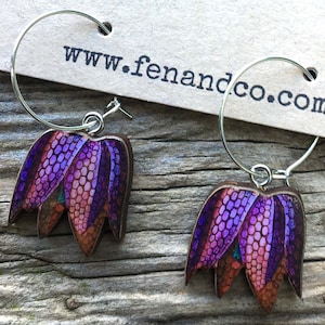 May include: A pair of silver hoop earrings with purple and orange flower charms. The charms are made of wood and have a detailed floral design. The earrings are from the website www.fenandco.com.