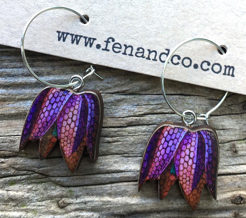 May include: A pair of silver hoop earrings with purple and orange flower charms. The charms are made of wood and have a detailed floral design. The earrings are from the website www.fenandco.com.