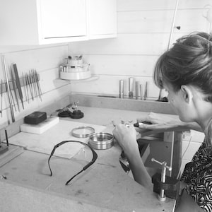 May include: A person working on jewelry in a workshop. The workspace is filled with tools and supplies, including a variety of files, a small anvil, and a pair of safety glasses.