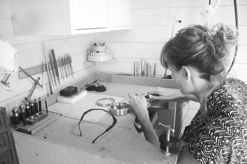 May include: A person working on jewelry in a workshop. The workspace is filled with tools and supplies, including a variety of files, a small anvil, and a pair of safety glasses.