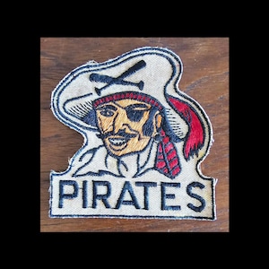 May include: A vintage embroidered patch featuring a cartoon pirate with a black eye patch, a red bandana, and a crossed swords design. The patch is beige with black and red embroidery and reads "PIRATES" in black lettering.