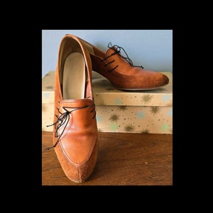 May include: A pair of brown leather and suede heeled shoes with black laces. The shoes are sitting on a box with a star pattern.