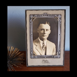 May include: A vintage black and white photograph of a man in a suit and tie. The photo is framed in a decorative frame with a floral design. The frame is made of cardboard and has a light brown color. The text "Payton Studio, Colorado Springs" is printed on the bottom of the frame.