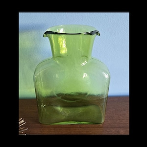 May include: A green glass vase with a square base and a flared, rounded top. The vase has a light green hue and sits on a wooden surface. The background is a light blue wall. The vase is empty.