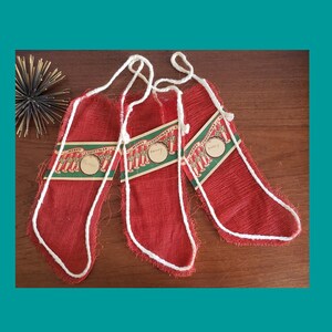May include: Three red burlap Christmas stockings with white trim and hanging loops. Each stocking has a decorative tag with the words "Merry Christmas" and a name. The stockings are on a wooden surface.