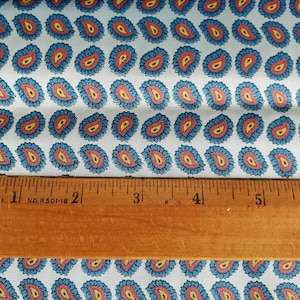 May include: White fabric with a repeating pattern of blue, red, and yellow paisley shapes. A wooden ruler with the text "MADE IN U.S.A. 1 NO. R 501-18 2 3 4 5 6" is placed on top of the fabric.