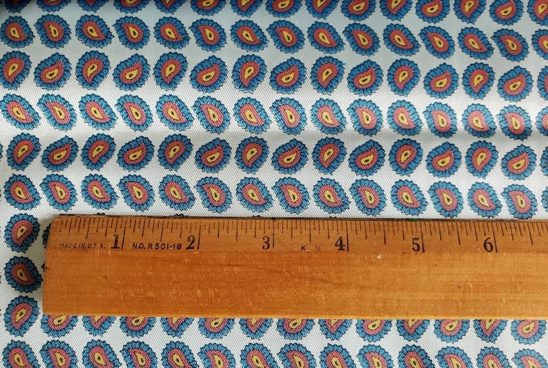 May include: White fabric with a repeating pattern of blue, red, and yellow paisley shapes. A wooden ruler with the text "MADE IN U.S.A. 1 NO. R 501-18 2 3 4 5 6" is placed on top of the fabric.