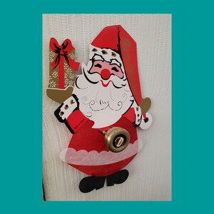 May include: A vintage Santa Claus door decoration. The red and white Santa figure has a gold-colored gift and a red hat with gold lettering. The decoration is attached to a door knob.