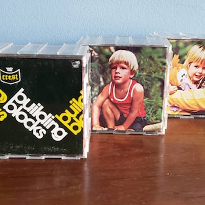 May include: Three clear plastic boxes with black and yellow building blocks packaging on the left box and photos of children on the right two boxes. The text on the left box reads "Crest Building Blocks".