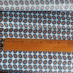 May include: A white fabric with a repeating pattern of blue and red paisley shapes. A wooden ruler is placed on top of the fabric. The ruler is marked with inches.