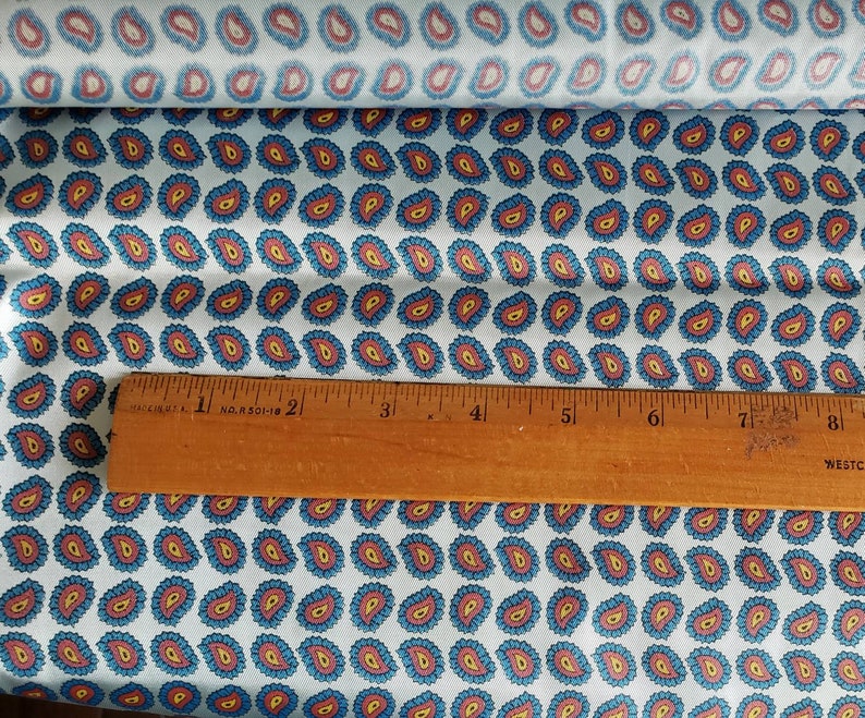 May include: A white fabric with a repeating pattern of blue and red paisley shapes. A wooden ruler is placed on top of the fabric. The ruler is marked with inches.