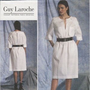 May include: A white dress with a V-neckline and a black belt. The dress has a pocket on the left side and is shown on a model with black heels. The pattern number is V1400 and the size range is 8-10-12-14-16.