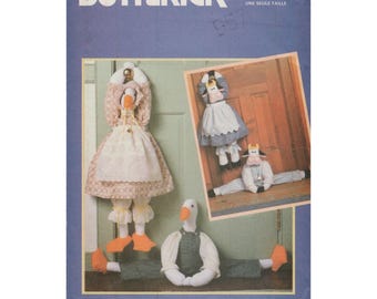 May include: A Butterick sewing pattern featuring a goose and cow door stop design. The goose door stop is wearing a dress and apron. The cow door stop is wearing a dress and a shirt. The pattern is designed by Wendy Everett.