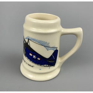 以下が含まれることがあります： 白い陶器製のビアジョッキに、青と黒の戦闘機のイラストが描かれています。飛行機の翼には白い星があり、下部には「VF-3 "Hellcat"」の文字が書かれています。