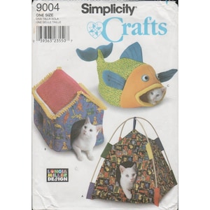 May include: Simplicity Crafts pattern 9004 featuring three pet bed designs: a house-shaped bed with a red roof, a fish-shaped bed with a green spotted body and orange face, and a tent-style bed with a patterned fabric, each occupied by a white cat.