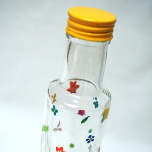 Puede incluir: Una botella de vidrio transparente con una tapa de metal amarilla. La botella está decorada con flores coloridas pintadas a mano y otras formas pequeñas. La botella es probablemente para almacenar líquidos.