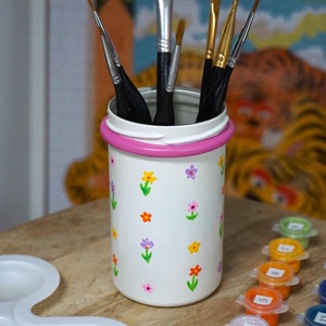 May include: A white painted jar with a pink band and floral designs holds a variety of paint brushes. The jar is sitting on a wooden surface with a white paint palette and small paint pots with numbers on them.