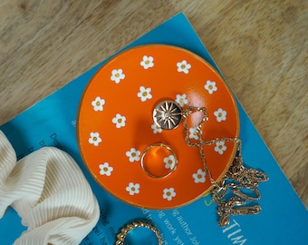 May include: Orange ceramic trinket dish with white daisy pattern, featuring a gold necklace with a sun pendant and a gold ring.