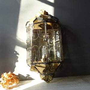 May include: A gold-toned metal wall sconce with a decorative, ornate design. The sconce has a clear glass shade with a geometric pattern and a single light bulb.