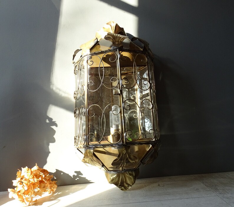 May include: A gold-toned metal wall sconce with a decorative, ornate design. The sconce has a clear glass shade with a geometric pattern and a single light bulb.