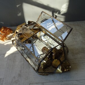 May include: A gold-toned metal and glass decorative box with a hinged lid. The box is decorated with ornate metalwork and has a geometric shape. The box is open and the lid is propped up.