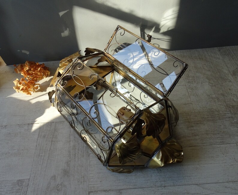 May include: A gold-toned metal and glass decorative box with a hinged lid. The box is decorated with ornate metalwork and has a geometric shape. The box is open and the lid is propped up.