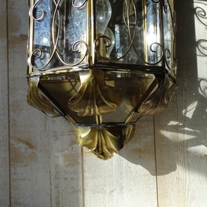 May include: A gold-toned metal lantern with a glass pane and intricate scrollwork design. The lantern has a decorative, ornate bottom with leaf-like details.