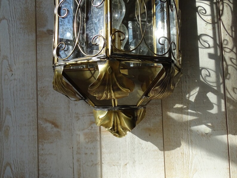 May include: A gold-toned metal lantern with a glass pane and intricate scrollwork design. The lantern has a decorative, ornate bottom with leaf-like details.