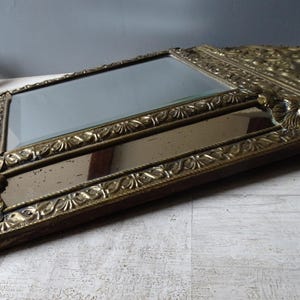 Puede incluir: Un espejo antiguo con un marco dorado ornamentado. El marco presenta dise&ntilde;os detallados de flores y volutas. El espejo tiene forma rectangular con otro espejo rectangular m&aacute;s peque&ntilde;o debajo. El espejo est&aacute; sobre una superficie clara.