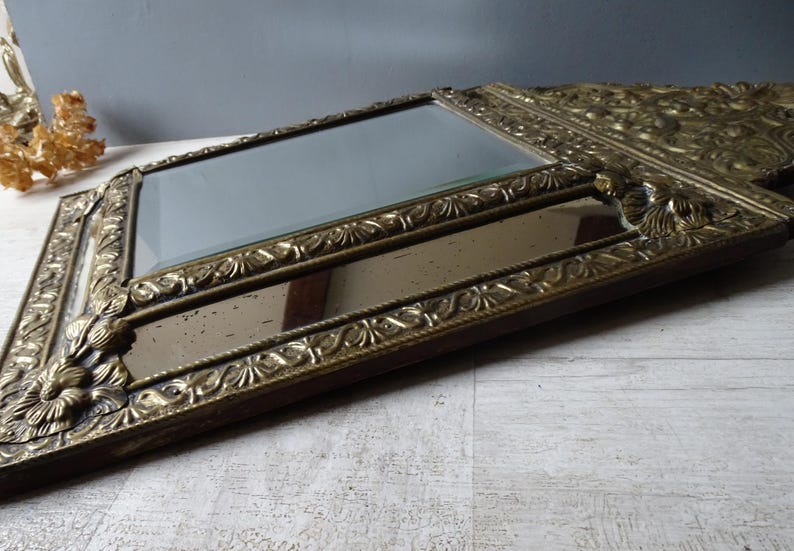 Puede incluir: Un espejo antiguo con un marco dorado ornamentado. El marco presenta dise&ntilde;os detallados de flores y volutas. El espejo tiene forma rectangular con otro espejo rectangular m&aacute;s peque&ntilde;o debajo. El espejo est&aacute; sobre una superficie clara.