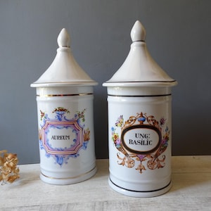 Könnte beinhalten: Zwei weiße Apothekergläser aus Keramik mit konischen Deckeln. Jedes Glas hat ein dekoratives Etikett mit floralen Akzenten und goldenen Verzierungen. Eines trägt die Aufschrift "AUREUM", das andere "UNG: BASILIC".