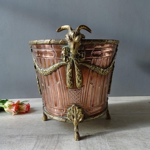 May include: A copper-toned metal planter with brass accents. The planter features a goat head handle, decorative garland, and ornate legs. The design includes a basket-like pattern and a bow. The planter is approximately 12 inches tall.