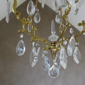 Puede incluir: Primer plano de una l&aacute;mpara de ara&ntilde;a dorada con m&uacute;ltiples colgantes de cristal transparente en forma de l&aacute;grima. La l&aacute;mpara tiene un dise&ntilde;o decorativo y ornamentado con brazos curvos y una columna central. Fondo de pared clara.