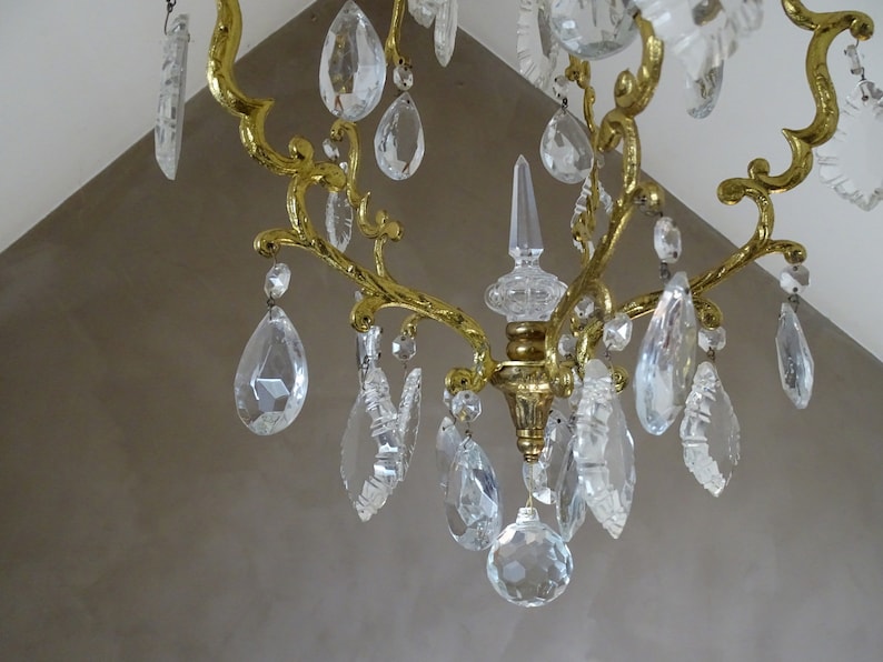 Puede incluir: Primer plano de una l&aacute;mpara de ara&ntilde;a dorada con m&uacute;ltiples colgantes de cristal transparente en forma de l&aacute;grima. La l&aacute;mpara tiene un dise&ntilde;o decorativo y ornamentado con brazos curvos y una columna central. Fondo de pared clara.