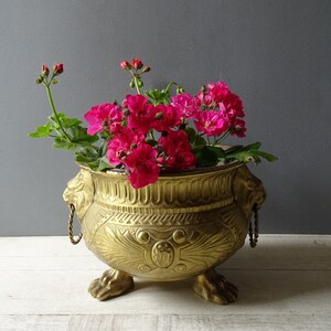 May include: A gold brass planter with lion head handles and feet. The planter is filled with pink flowers.