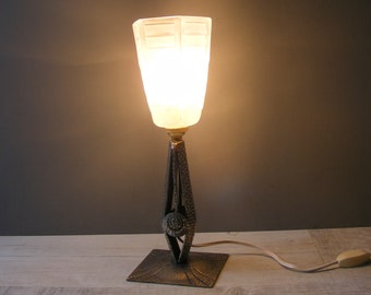 May include: A vintage table lamp with a frosted glass shade and a wrought iron base. The base features a decorative design with a circular motif. The lamp is lit and casting a warm glow.