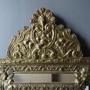 Puede incluir: Espejo dorado ornamentado con intrincados detalles florales y de volutas. El espejo rectangular tiene una parte superior decorativa con un dise&ntilde;o central y detalles florales en los lados. El espejo est&aacute; sobre un fondo gris.