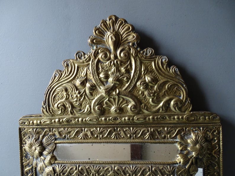 Puede incluir: Espejo dorado ornamentado con intrincados detalles florales y de volutas. El espejo rectangular tiene una parte superior decorativa con un dise&ntilde;o central y detalles florales en los lados. El espejo est&aacute; sobre un fondo gris.