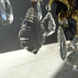 Puede incluir: Primer plano de una l&aacute;mpara de ara&ntilde;a vintage con colgantes de cristal transparente y herrajes de lat&oacute;n. Los cristales tienen diversas formas, incluyendo l&aacute;grimas y facetas, reflejando la luz. El lat&oacute;n presenta una p&aacute;tina c&aacute;lida y envejecida.