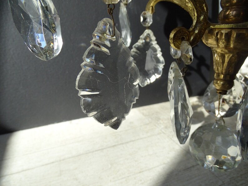 Puede incluir: Primer plano de una l&aacute;mpara de ara&ntilde;a vintage con colgantes de cristal transparente y herrajes de lat&oacute;n. Los cristales tienen diversas formas, incluyendo l&aacute;grimas y facetas, reflejando la luz. El lat&oacute;n presenta una p&aacute;tina c&aacute;lida y envejecida.