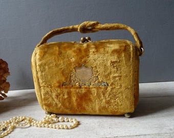 May include: A vintage mustard yellow velvet handbag with a gold metal clasp and a decorative shield design. The bag has a top handle and a chain strap.