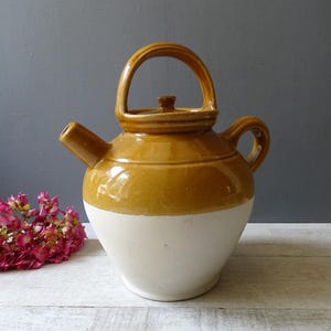 May include: A two-tone ceramic teapot with a handle, spout, and lid. The top half is a warm, mustard yellow, while the bottom half is white. The teapot sits on a white wooden surface, with pink flowers in the foreground.