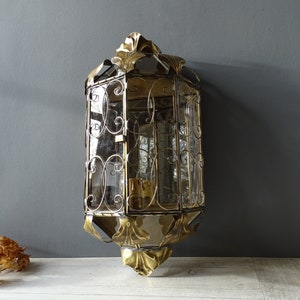 May include: A gold-toned metal wall sconce with a decorative, ornate design. The sconce has a glass panel and a clear glass shade.