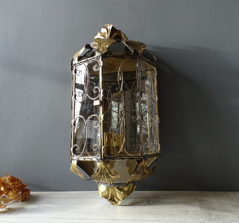 May include: A gold-toned metal wall sconce with a decorative, ornate design. The sconce has a glass panel and a clear glass shade.